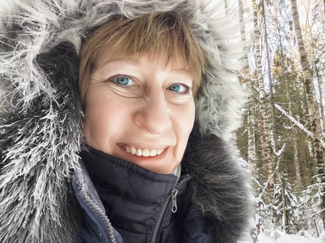A mature blonde woman takes a cheerful selfie in a snowy forest landscape, dressed warmly for the cold weather.
