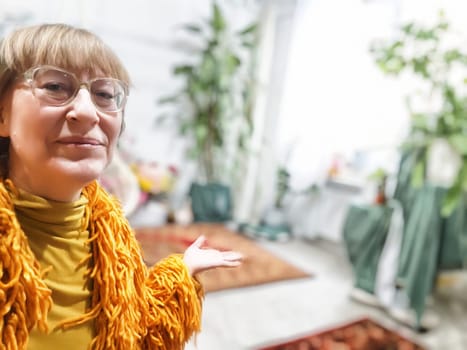 A mature blonde woman smiles while gesturing towards her colorful living room, sharing her lifestyle with followers.