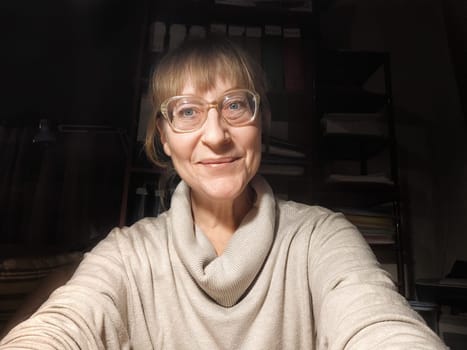 A blonde middle-aged blogger poses for a fun selfie in her cozy workspace, reflecting her lifestyle and cheerful personality.
