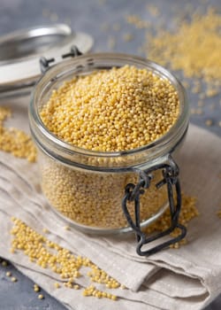 Glass jar of raw uncooked yellow millet grain on napkin on gray table closeup. Ancient pseudocereal seeds. Organic super food. Healthy food concept. Gluten free alternative seeds
