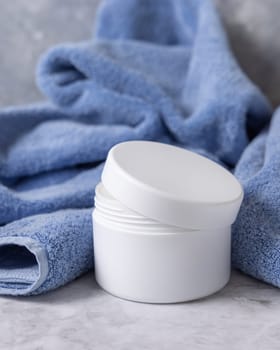 White opened cream jar with blank lid near blue bath towel close up, brand packaging mockup. Natural herbal organic cosmetics for daily facecare and spa procedures, copy space