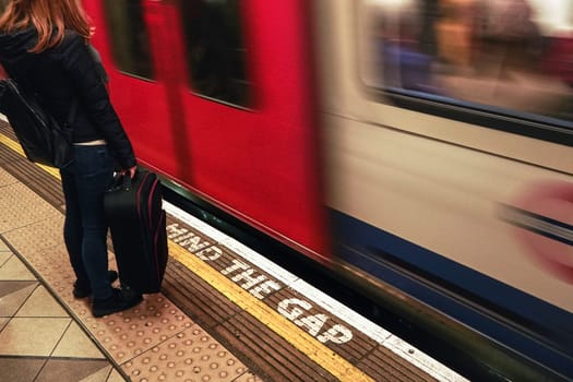 London, United Kingdom - February 01, 2019: Girl with her black suitcase waiting at London underground station, as train is arriving behind yellow line with Mind the Gap warning