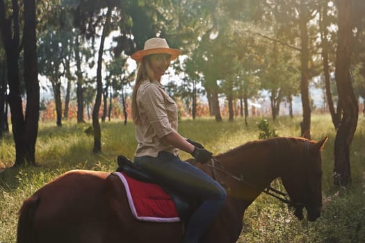 Young woman in shirt and straw hat, riding brown horse in the park, blurred grass and trees background