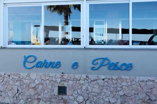 Catania. Italy - February 24, 2025:  Vibrant Sicilian seaside restaurant with a Carne e Pesce sign, large windows reflecting palm trees, and diners enjoying ocean views, perfect for food and travel themes.