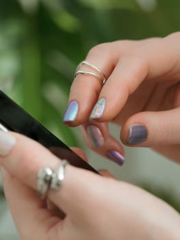 Close-up of hands with colorful nail polish using Phone. female hands with beautiful rings and skittles nails use smartphone on green foliage monstera background. Vertical