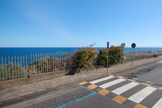 Coastal pedestrian safety. A pedestrian crossing sign stands by a scenic coastal road in Catania, Sicily, Italy, with vibrant flowers and the sea in the background.