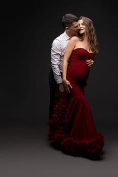 Tender man embracing pregnant woman in red elegant dress while standing on gray background in studio and kissing