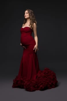 Serene pregnant female wearing long red dress standing with closed eyes on gray background and touching tummy