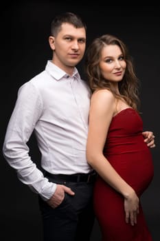 Handsome young man in classy outfit hugging beautiful pregnant wife in elegant red dress and looking at camera while standing against black background