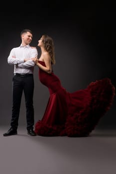 Full body of charming young pregnant female in elegant red gown standing near confident husband in studio with gray background