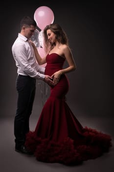 Side view of happy young man in elegant outfit touching belly from pregnant wife - stylish dress while standing together - dark studio decorated with pink balloons