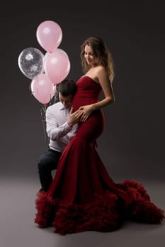 Side view of happy young man in elegant outfit hear belly from pregnant wife - stylish dress while standing together - dark studio decorated with pink balloons