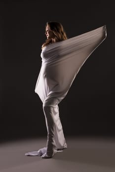 Sensual nude pregnant female wrapped in white cloth standing on dark background in studio
