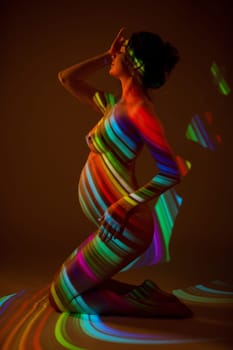 Side view of sensual nude pregnant female standing on knees in dark studio with colorful neon illumination