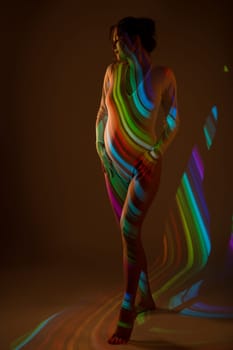 Graceful pregnant female in white bodysuit standing in dark studio illuminated by striped color light of projector