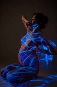 Side view of naked pregnant female sitting with closed eyes on floor in dark studio with blue neon light
