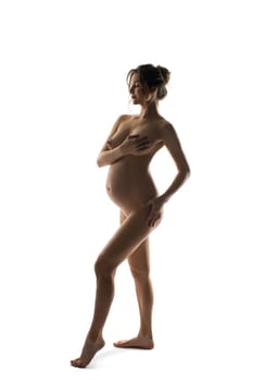 Side view of happy healthy tender pregnant female with belly covering breast with hands while standing against white background