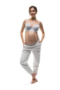 Full body of barefoot young pregnant female in white bra and pants smiling and looking at camera against white background