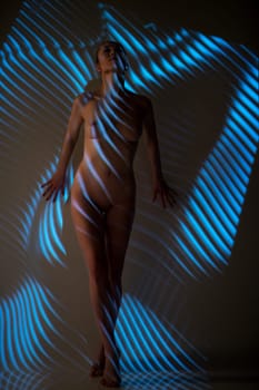 Full body of dreamy young naked female model standing near wall in dark studio with blue striped illumination