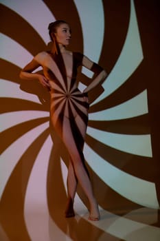 Young slim nude female model posing under light of projector creating geometric striped ornament on body and wall in studio