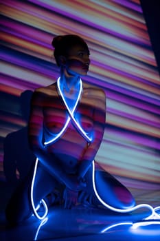 Unrecognizable slim naked female model with glowing neon strap wrapped around body sitting under light of projector creating colorful striped pattern in studio