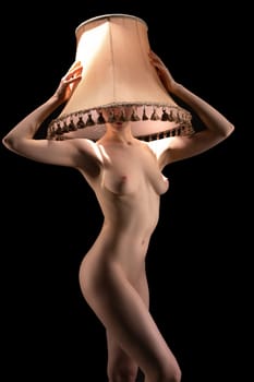 Slim curvy naked female model covering head and face with beige lampshade on black background
