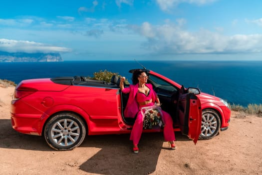 A woman is sitting in the back of a red convertible car. The car is parked on a dirt road near the ocean. The woman is wearing a pink suit and red shoes. Scene is relaxed and carefree