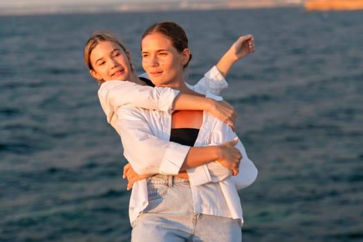 A woman in a white shirt and jeans holds a girl in a white shirt in her arms. They are both smiling and looking at the camera. The background is a body of water.