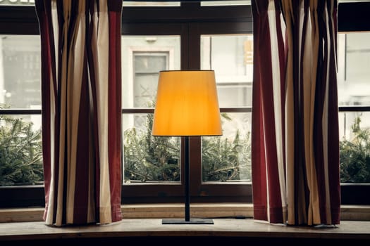 A warmly lit table lamp on a rustic windowsill, surrounded by striped curtains and greenery outside. A perfect composition for interior, decor, and cozy lifestyle projects