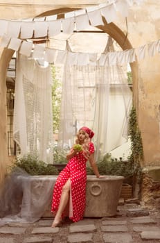 A young blonde woman is sitting in the courtyard of the old city, holding grapes in her hands. She is wearing a red dress with white polka dots and a headscarf.