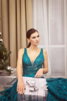 A beautiful girl in an evening blue dress sits against the background of a Christmas tree and holds a box with a gift. The girl is elegant, with a hairstyle and makeup. The girl is dreaming.