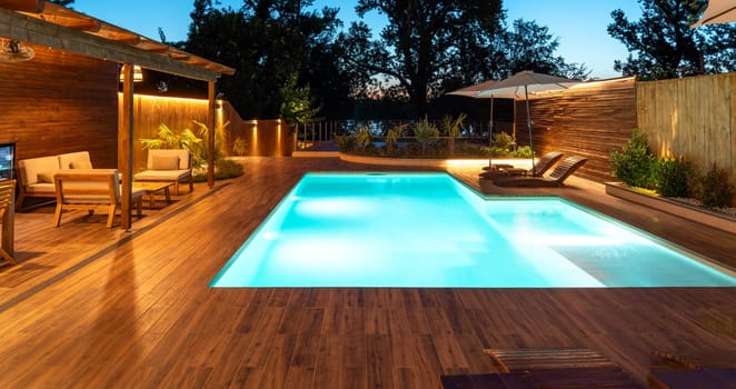 Luxurious backyard pool with a wooden deck and cozy lounge area. Evening retreat illuminated with warm, inviting lighting