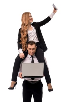 Businesswoman in formal wear doing selfie on smartphone while sitting on shoulders of businessman using laptop isolated on white background