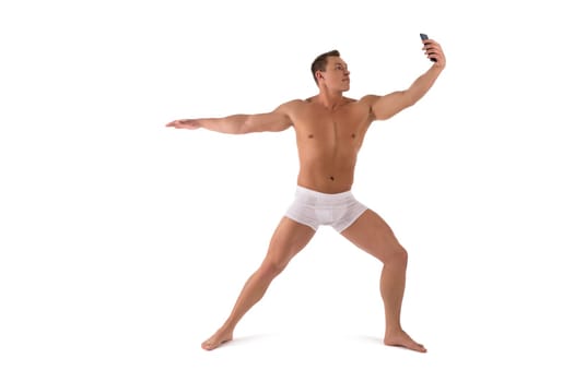 Full body of confident athletic male in white underpants taking selfie on mobile phone while standing in Warrior pose against white background