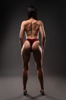 Full body of anonymous topless female with muscular back wearing thong and high heels against gray background