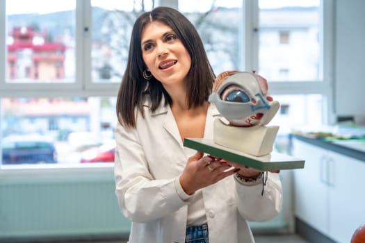 Young woman wearing lab coat holding anatomical model of eye and optic nerve, explaining its function in university classroom