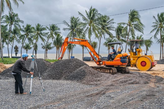 Construction heavy machinery excavator and builder with theodolite at a construction site on a beach by the sea in Asia. Nha Trang, Vietnam - December 14, 2024