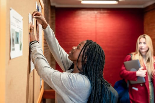 Focused student pinning a paper on a bulletin board in a university hallway, with another student walking in the background