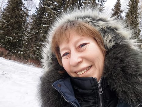 A middle-aged blonde woman stands in a snowy forest, smiling brightly as she captures a selfie during her winter trip.