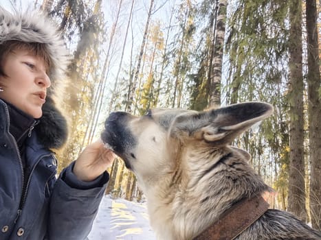 A middle-aged woman takes a selfie with her German Shepherd during a chilly winter outing in a snowy forest.