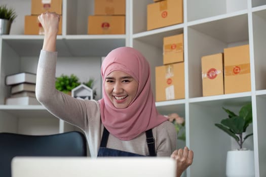 A joyful Muslim woman in a hijab raises her fist in celebration, marking a successful moment in her online business journey.