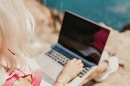 A woman is sitting on a rock with a laptop open in front of her. She is typing on the keyboard and she is focused on her work. Concept of productivity and concentration