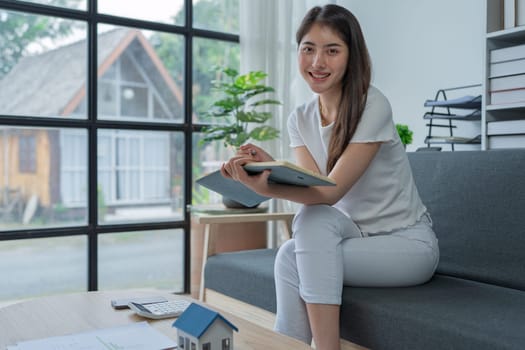 Young woman analyzing financial documents at home with calculator and model house on table in modern living room.