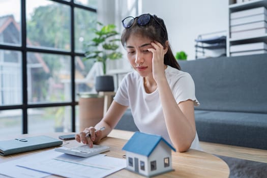 Young woman analyzing financial documents at home with calculator and model house on table in modern living room.