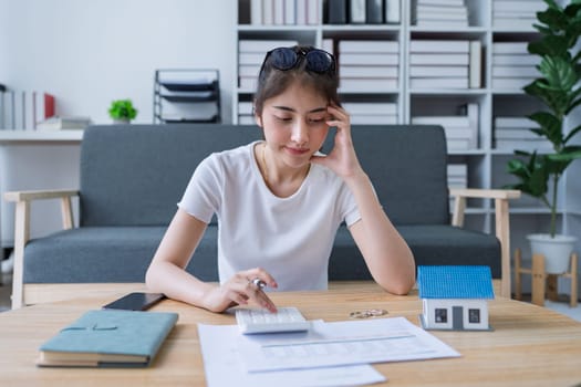 Young woman analyzing financial documents at home with calculator and model house on table in modern living room.