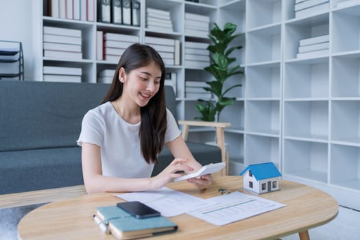 Young woman analyzing financial documents at home with calculator and model house on table in modern living room.