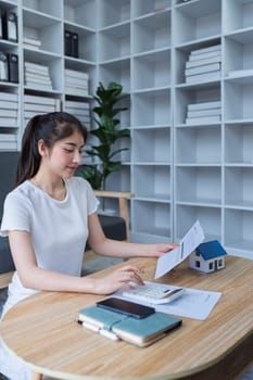 Young woman analyzing financial documents at home with calculator and model house on table in modern living room.