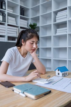 Young woman analyzing financial documents at home with calculator and model house on table in modern living room.