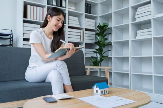 Young woman analyzing financial documents at home with calculator and model house on table in modern living room.