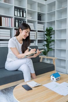 Young woman analyzing financial documents at home with calculator and model house on table in modern living room.